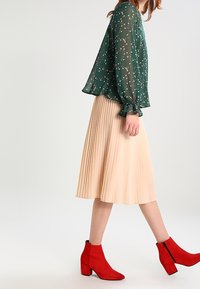 Blouse en chiffon à fleurs vertes avec des manches bouffantes, associée à une jupe plissée beige et des bottines rouges à talons carrés.