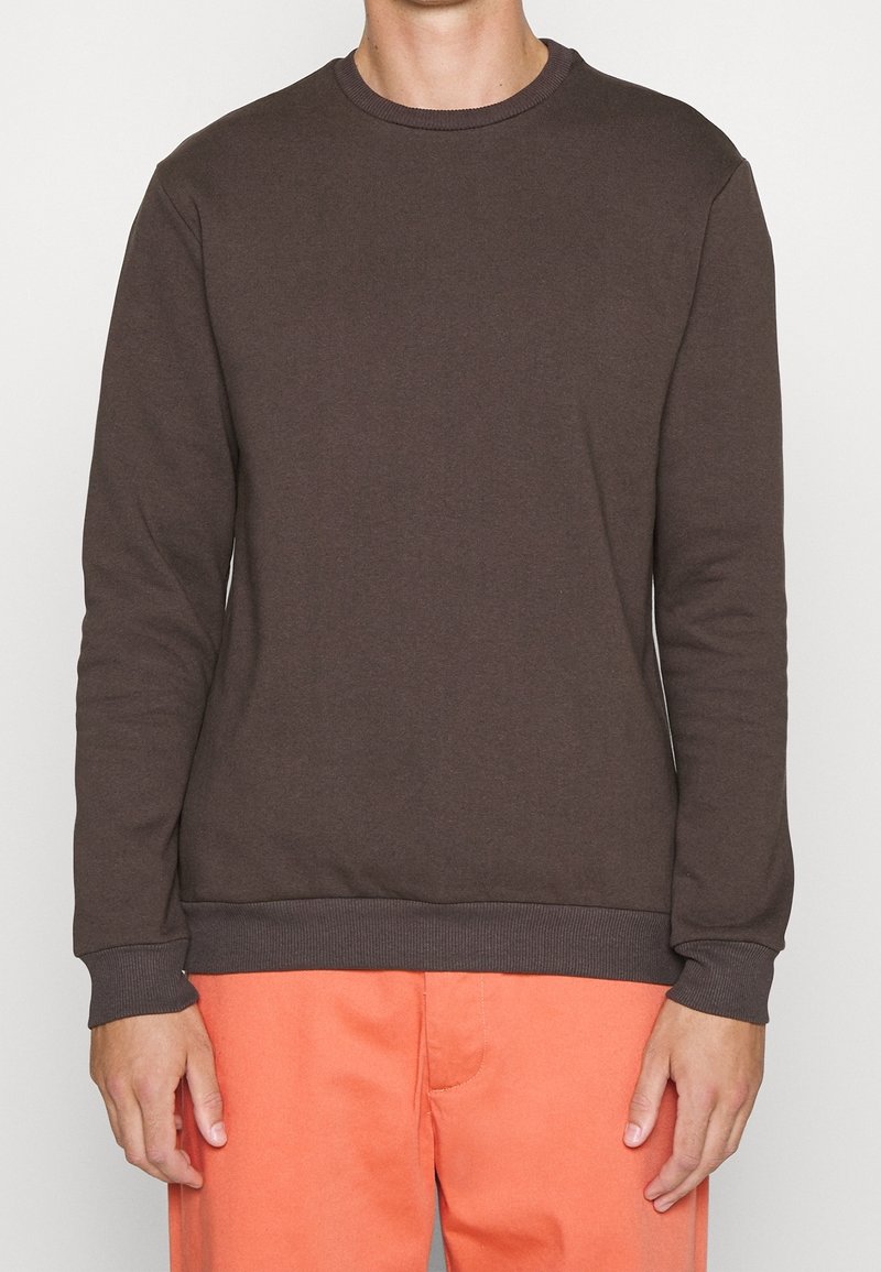 Personne portant un sweat-shirt à col rond marron foncé uni et un pantalon orange vif, debout devant un fond blanc uni.