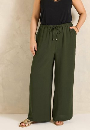 Kvinde iført olivengrønne bukser med vide ben og bindebånd i taljen, sort ærmeløs top og guld flade sandaler foran en beige baggrund.