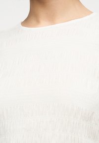 Κοντινό πλάνο ατόμου που φορά λευκή μπλούζα από υφαντό ύφασμα με μαζεμένες, ρυχωτές λεπτομέρειες και στρογγυλή λαιμόκοψη.