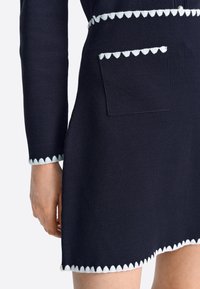 Tumesinine kleit tekstureeritud kootud kangast. Omab valget lainelist äärist, väikest taskut esiküljel ja keha järgivat lõiget.