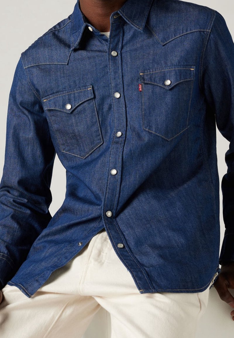 Homme portant une chemise en jean bleu foncé à boutons-pression avec poches, associée à un pantalon blanc cassé.