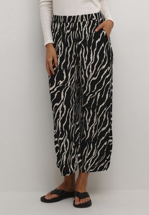 Femme portant un pantalon large imprimé zèbre noir et blanc, un haut blanc à manches longues et des sandales noires, debout avec une main dans la poche.