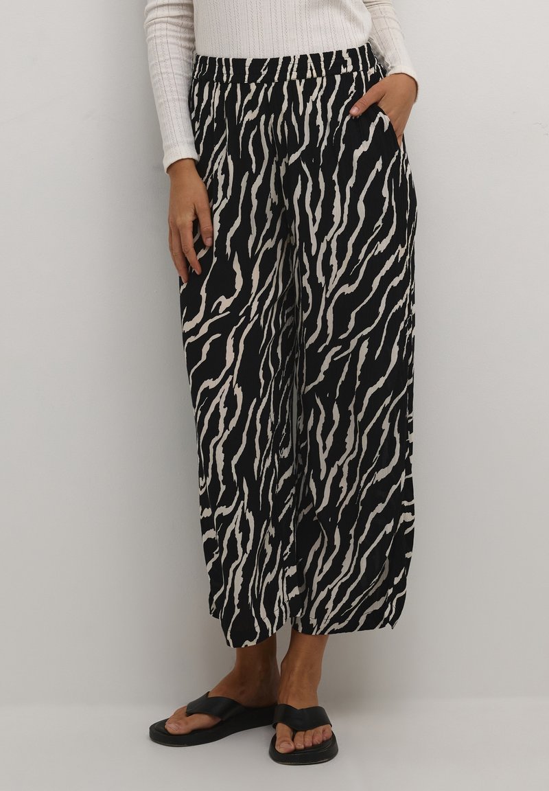 Femme portant un pantalon large imprimé zèbre noir et blanc, un haut blanc à manches longues et des sandales noires, debout avec une main dans la poche.