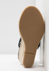 Chaussure à talon compensé avec semelle en caoutchouc beige lisse, plateforme style espadrille tissée, et bride noire à la cheville posée contre une surface blanche.