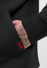 Mano con tatuaggio parzialmente all'interno di tasca di un hoodie nero con un'etichetta rossa "UFC" sul bordo della tasca.