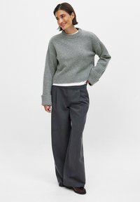 Grå strikket sweater med ribbede nederkant, parret med brede mørkegrå bukser. Sættet er sammensat med en lagdelt hvid skjorte under.