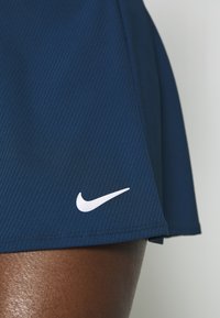 Sportovní skort tmavě modré barvy z žebrované látky, s bílým logem Nike na lemu, s hladkým a texturovaným povrchem.