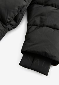 Melnā uzpūsto jaku ar quilted tekstūru. Ir saliekama piedurkne, kas atklāj zīmola elastīgas aproces. Materiāls izskatās spīdīgs un gluds.