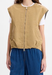 Tarnfarbende ärmellose Jacke mit hohem Kragen, Frontreißverschluss und zwei Seitentaschen. Sie hat eine glatte Textur und eine lockere Passform. Wird über einem weißen Hemd getragen.