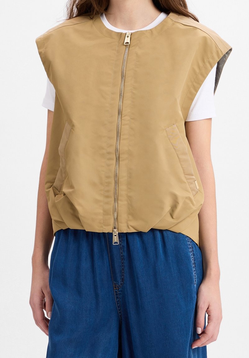 Tarnfarbende ärmellose Jacke mit hohem Kragen, Frontreißverschluss und zwei Seitentaschen. Sie hat eine glatte Textur und eine lockere Passform. Wird über einem weißen Hemd getragen.