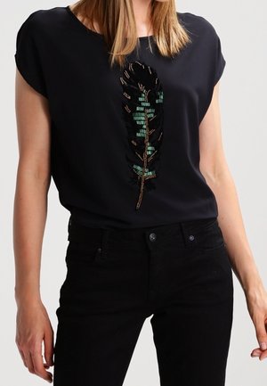 Femme portant un haut noir à manches courtes avec un motif plume orné de perles et un pantalon noir, debout les bras détendus le long du corps.