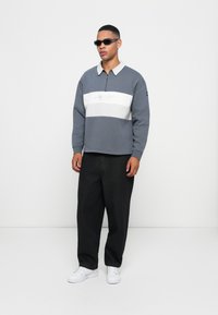Pullover cinza com riscas horizontais brancas, gola com zíper de um quarto e logótipo bordado; combinado com calças largas pretas e sapatilhas brancas.