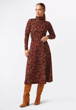Abito lungo color marsala con maniche lunghe e collo alto, caratterizzato da una gonna fluida e un motivo astratto giallo e verde; abbinato a stivali marroni alti fino al ginocchio.