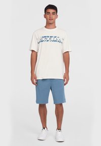 Lys beige t-shirt med blåt grafisk design, parret med blå shorts. Outfitet har en afslappet pasform og en casual stil. Hvide sneakers fuldender looket.