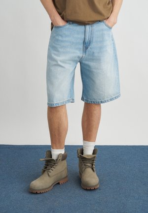 Άτομο που φοράει ανοιχτό μπλε τζιν σορτς, καφέ πουκάμισο, λευκές κάλτσες και μπεζ μποτάκια με κορδόνια, στέκεται σε μπλε χαλί.