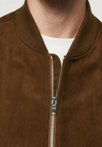 Chaqueta bomber de ante marrón con cuello acanalado, cierre frontal con cremallera y textura suave, con detalles en hardware plateado.