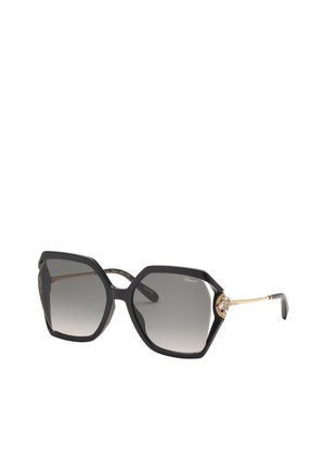 Lunettes de soleil hexagonales noires avec branches dorées, verres gris dégradés et accents dorés décoratifs près des charnières.