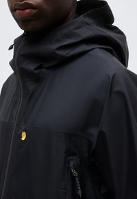 Bărbat purtând o jachetă neagră impermeabilă cu glugă, buzunar cu fermoar și un mic logo auriu pe piept.