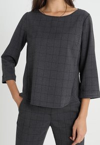 Blusa grigia a quadri con scollatura a barca e maniche a tre quarti, realizzata in un tessuto strutturato, caratterizzata da un motivo a quadri.