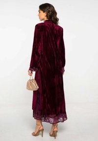 Long manteau en velours bordeaux avec une finition texturée, bordure en dentelle semi-transparente aux manches, associé à un sac en satin doré et des talons à lanières.