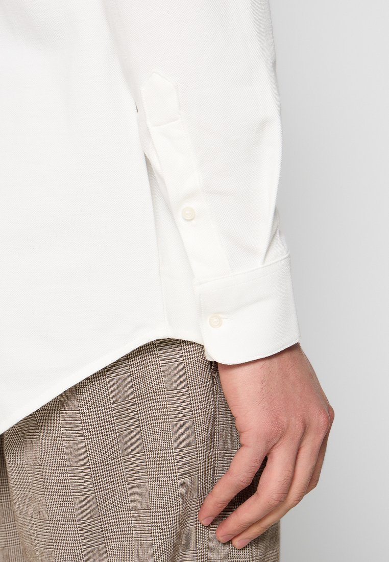 Biała koszula z długim rękawem o teksturowanym splocie, z mankietami zapinanymi na guziki. W komplecie z brązowymi spodniami w kratkę.