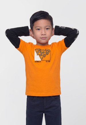 Un garçon portant un tee-shirt orange à motif graphique et un pantalon foncé se tient debout, les mains derrière la tête, sur un fond blanc uni.