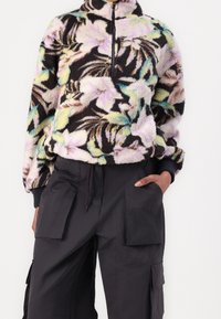 Polarowy pulower z kwiatowym wzorem w pastelowych kolorach na czarnym tle, z półzamkiem i przednią kieszenią. Połączony z czarnymi bojówkami.