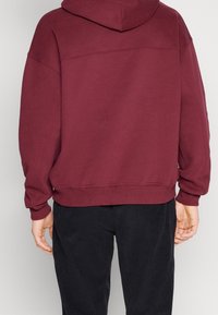 Sudadera con capucha en color burdeos, de corte relajado, con puños y dobladillo acanalados, que presenta una textura suave y costuras en la parte posterior.