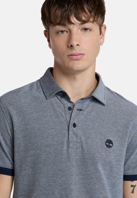 Harmaa lyhythihainen polo paita, jossa on teksturoitu kangas, kaksinappinen napitus, sävy sävyyn kaulus ja pieni logo rinnassa. Laivastonsinisiä yksityiskohtia hihoissa.