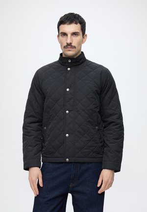 Mand med mørkt hår og overskæg iført sort quiltet jakke med trykknapper og mørkeblå jeans, står mod en ensfarvet lys baggrund.