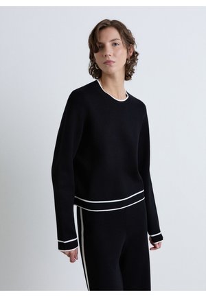 Young person with short curly hair wears black long-sleeve top and pants with white trim, standing against plain light gray background.