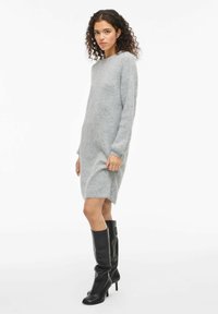 Hellgraues Strickkleid mit langen Ärmeln, Rundhalsausschnitt und seitlichen Schlitzen, kombiniert mit knielangen schwarzen Lederstiefeln mit Absatz.