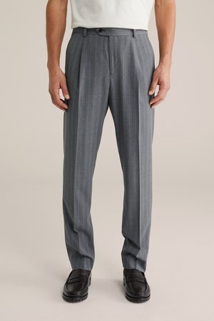 Vyras dėvi pilkus dryžuotus kelnes ir juodus odinius loaferius, stovintis atsipalaidavęs su rankomis šalia kūno, priešais neutralią foną.