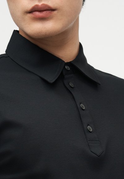 Κοντινό πλάνο ενός ατόμου που φοράει μαύρο πόλο με κουμπιά, εμφανίζοντας το γιακά και την περιοχή του άνω στήθους.