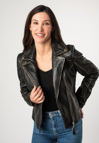 Zwarte leren motorjack met een gesneden kraag, asymmetrische rits, tonale stiksels en zijzakken. Model draagt een strakke spijkerbroek.
