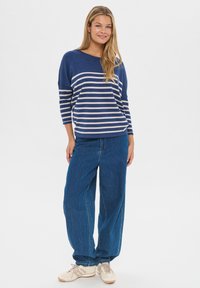 Jeune femme debout et souriante, portant un pull rayé bleu, un jean bleu ample et des baskets beiges sur un fond uni.