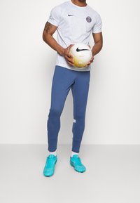 Camiseta blanca de Nike con estampado y pantalones deportivos azules, sosteniendo un balón de fútbol blanco y dorado. Zapatillas deportivas azules completan el atuendo.