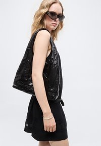 Γυναίκα με κυματιστά ξανθά μαλλιά φοράει μαύρο αμάνικο φόρεμα, μεγάλο γυαλιστερό μαύρο τσάντα ώμου, σκούρα γωνιώδη γυαλιά ηλίου και ασημένιο βραχιόλι.