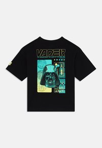 Svart bomull t-shirt med ett grafiskt tryck av en hjälmad figur och text i neongrön och gult, med texturer och former kopplade till rymden.