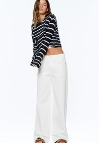 Femme portant un pull court rayé noir et blanc à manches longues et un pantalon blanc taille haute à jambes larges devant un fond uni.