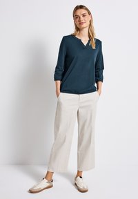 Teal Longsleeve-Oberteil mit V-Ausschnitt und elastischen Bündchen, kombiniert mit cremefarbenen weiten Hosen und neutralen Sneakers. Glattes Material mit minimalistischem Design.