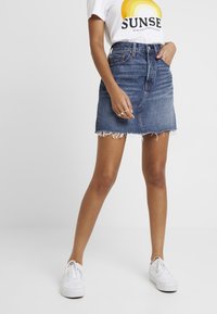 Jupe en denim avec un ourlet effiloché, lavage bleu, taille haute et poches avant, assortie à un t-shirt graphique blanc et des baskets blanches.