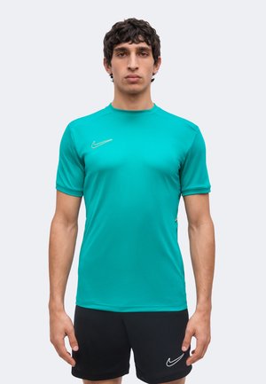 Jeune homme debout face à l'avant, portant un t-shirt de sport turquoise à manches courtes et un short noir avec des logos Nike blancs.