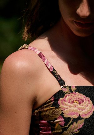 Woman wearing a floral dress with thin straps, sunlight casting shadows on her shoulder and part of her face.