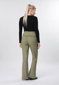 Ξανθιά γυναίκα με αλογοουρά, ντυμένη με μαύρο μακρυμάνικο τοπ και πράσινο πλεκτό παντελόνι με πτυχώσεις, στέκεται μπροστά σε απλό λευκό φόντο.