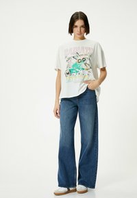 Hvid grafisk t-shirt med farverige tegneserie designs, parret med brede blå denim jeans og hvide sneakers med brune såler.