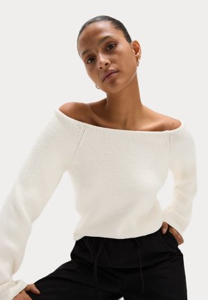 Pull en tricot blanc, décolleté épaule, avec de longues manches, texture côtelée et design ajusté, associé à un pantalon noir à cordon de serrage.