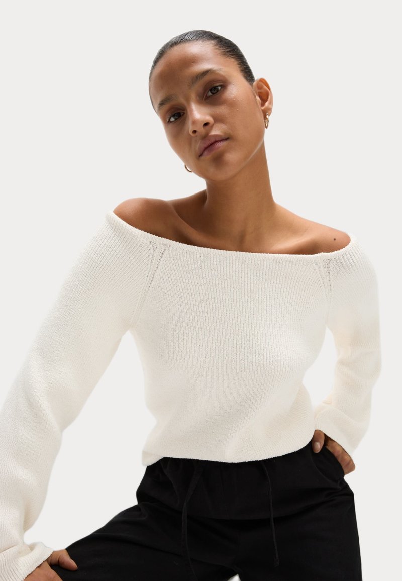 Vit, off-shoulder stickad tröja med långa ärmar, ribbad textur och figurnära design, tillsammans med svarta byxor med dragsko.