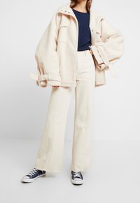 Överdimensionerad krämfärgad ulljacka med tryckknappar, tillsammans med högmidjade krämfärgade vida jeans och marinblå canvas sneakers. Ribbade marinblå topp under.